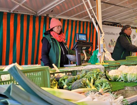 Zwei Verkäufer an einem Gemüsestand auf einem Wochenmarkt mit Brokkoli und Blumenkohl im Vordergrund.