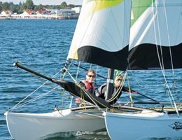 Zwei Personen segeln auf einem Katamaran auf einem See mit Küste im Hintergrund.