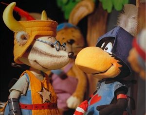 Zwei Puppenfiguren in Ritterkostümen, eine Maus und ein Rabe, stehen nebeneinander. Im Hintergrund ist ein Hase zu sehen.