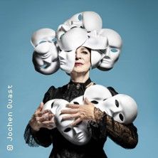 Eine Person in schwarzer Kleidung hält weiße Masken und trägt einen Kopfschmuck aus Masken vor blauem Hintergrund.