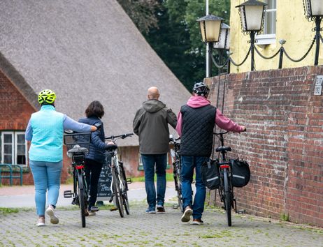 Vier Personen schieben Fahrräder entlang einer gepflasterten Gasse mit Backsteinmauern und einem Reetdachhaus im Hintergrund.