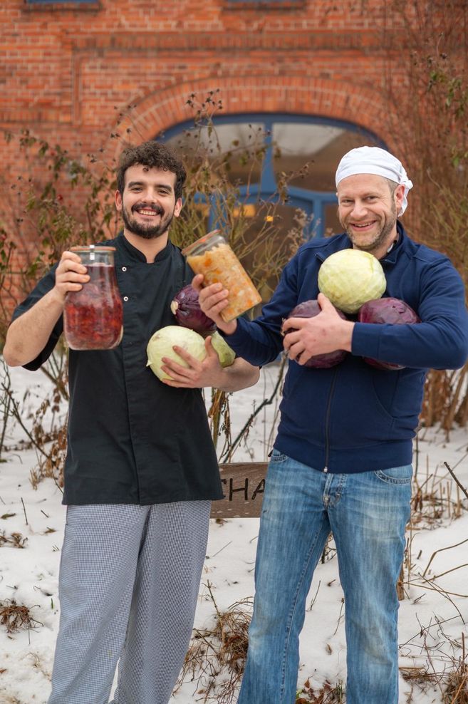 Zwei Männer halten Gläser mit fermentiertem Gemüse und Kohlköpfe in den Armen, vor einem Backsteingebäude im Schnee.