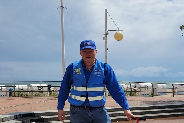 Ein Mann in blauer Kleidung und Warnweste steht vor einer Strandpromenade mit Strandkörben im Hintergrund.