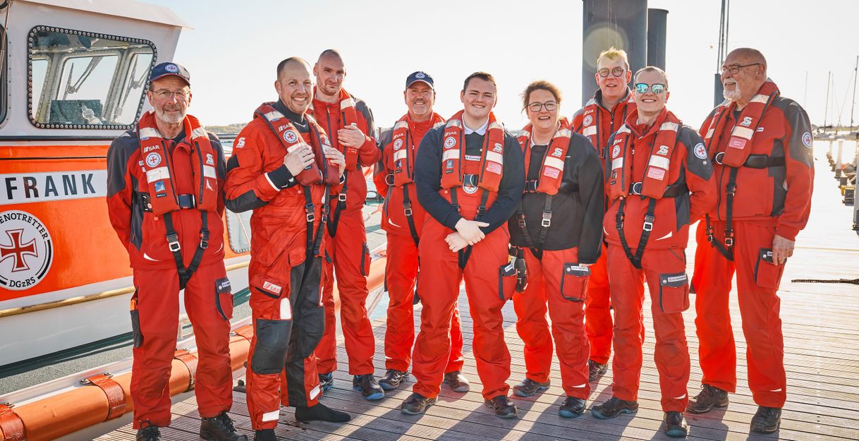 Gruppe von Menschen in roten Rettungsanzügen steht auf einem Steg vor einem Rettungsboot.