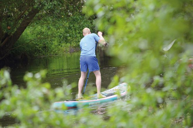 Person beim Stand-up Paddling auf einem Fluss, umgeben von grüner Vegetation.
