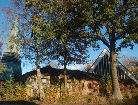 Kirche mit spitzem Turm und modernem Giebel, umgeben von Bäumen im Herbst.