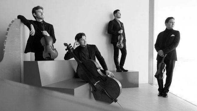 Vier Musiker in Anzügen posieren mit ihren Streichinstrumenten in einem modernen Raum.