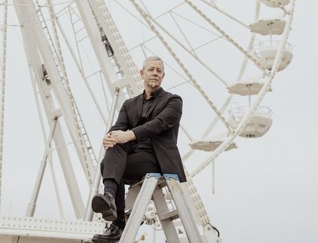 Eine Person in schwarzem Anzug sitzt auf einer Leiter vor einem Riesenrad.