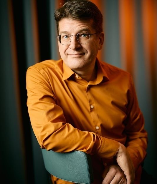 Person in orangefarbenem Hemd sitzt auf einem Stuhl vor einem gestreiften Hintergrund.