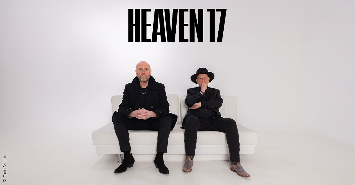 Zwei Männer sitzen auf einem weißen Sofa vor einem weißen Hintergrund mit dem Schriftzug 'Heaven 17' darüber.