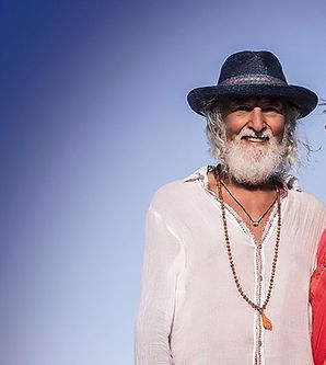 Ein Mann mit Hut und Bart steht neben einer Frau in rotem Kleid vor blauem Hintergrund.
