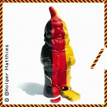 Ein Gartenzwerg, bemalt in den Farben der deutschen Flagge, steht auf einem weißen Hintergrund. Der Zwerg ist in der Mitte gespalten.