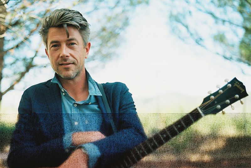 Ein Mann in einem blauen Anzug steht im Freien mit einer Gitarre im Vordergrund.