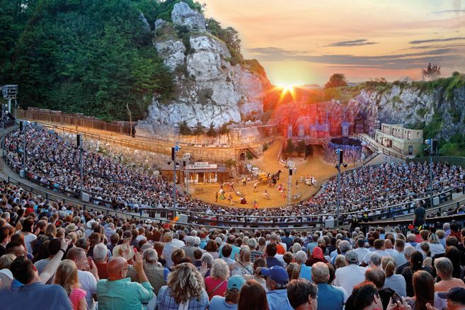 Eine große Menschenmenge sitzt in einem Freilufttheater, umgeben von felsigen Landschaften. Die Sonne geht im Hintergrund unter und taucht die Szene in warmes Licht.
