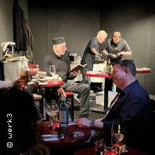 Eine Szene in einem Restaurant mit mehreren Personen, die an Tischen sitzen und essen. Im Hintergrund arbeiten zwei Köche.