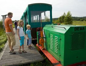 Eine Familie steht neben einer kleinen grünen Moorbahn mit der Aufschrift 'DIEMA' auf einem Holzsteg in einer grünen Landschaft.