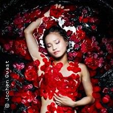 Eine Frau liegt in einem Wasserbecken, umgeben von roten Rosenblättern, mit geschlossenen Augen und entspanntem Gesichtsausdruck.