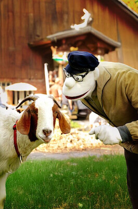 Eine Puppe in einer Jacke und Mütze beugt sich zu einer Ziege auf einer Wiese vor einem Holzgebäude.