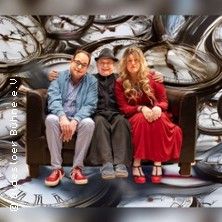 Drei Personen sitzen auf einem Sofa vor einem Hintergrund mit vielen Uhren.