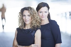 Zwei Frauen in eleganter Kleidung stehen nebeneinander und schauen in die Kamera.