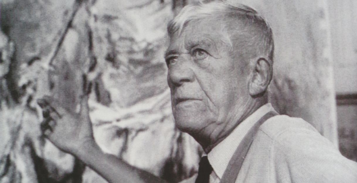 Ein älterer Mann steht vor einem großen Gemälde und gestikuliert mit der Hand.