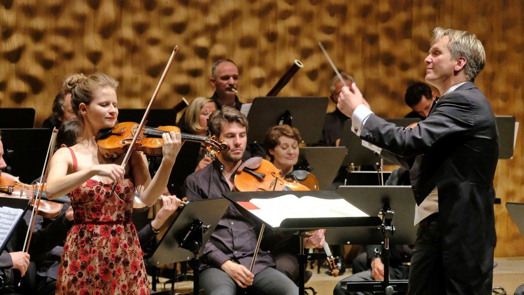 Eine Violinistin spielt vor einem Orchester, dirigiert von einem Mann im Anzug.