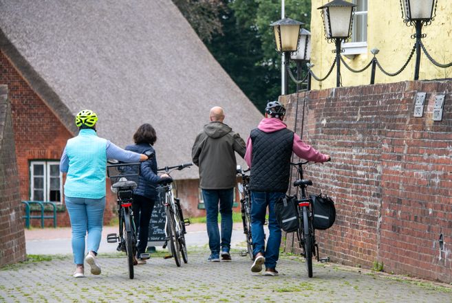 Vier Personen schieben Fahrräder entlang einer gepflasterten Gasse mit Backsteinmauern und einem Reetdachhaus im Hintergrund.