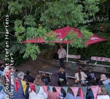 Ein Stand-up-Comedian tritt im Freien unter einem roten Sonnenschirm auf, umgeben von Bäumen und einem Publikum, das auf Bänken sitzt.