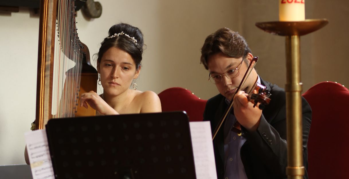 Zwei Musiker spielen Harfe und Geige in einem Raum mit einer großen Kerze.