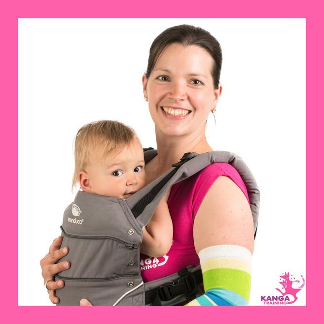 Frau mit Baby in einer Trage, lächelnd, vor weißem Hintergrund mit pinkem Rahmen und Kanga Training Logo.