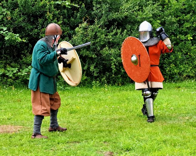 Two fighters in historical armor are practicing a battle on a meadow. One is carrying a shield and a sword, while the other is wearing armored armor and has a large shield.