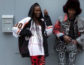 Zwei Menschen stehen modern gekleidet vor eine grauen Häuserwand.