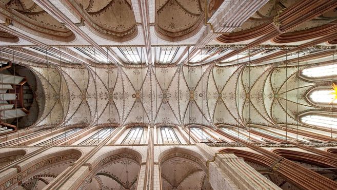 Blick auf die kunstvoll verzierte Decke einer gotischen Kathedrale mit hohen Fenstern und Orgelpfeifen.