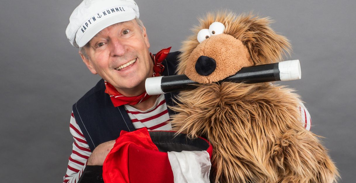 Ein Mann in Matrosenkleidung mit einer weißen Mütze und ein Plüschtier mit einem Zauberstab im Mund.