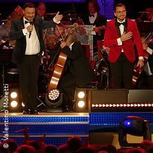 Zwei Männer in Anzügen auf einer Bühne mit Orchester im Hintergrund.