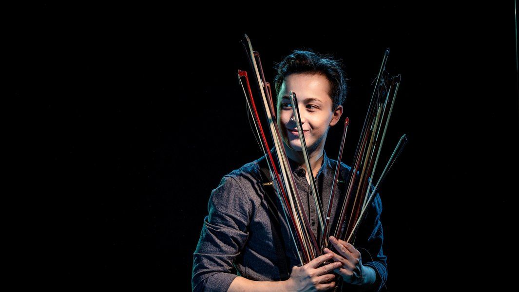 Person hält mehrere Geigenbögen vor schwarzem Hintergrund.