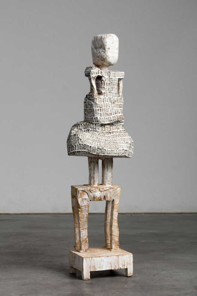 Eine abstrakte Skulptur aus Holz, die eine menschliche Figur darstellt. Sie hat einen runden Kopf und eine strukturierte Oberfläche.