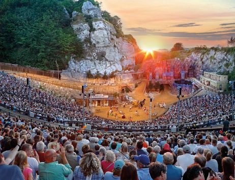Eine große Menschenmenge sitzt in einem Freilufttheater, umgeben von felsigen Landschaften. Die Sonne geht im Hintergrund unter und taucht die Szene in warmes Licht.