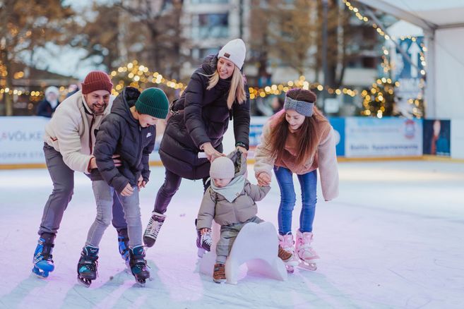 Eine Familie beim Schlittschuhlaufen auf einer Eisbahn, umgeben von Lichterketten.