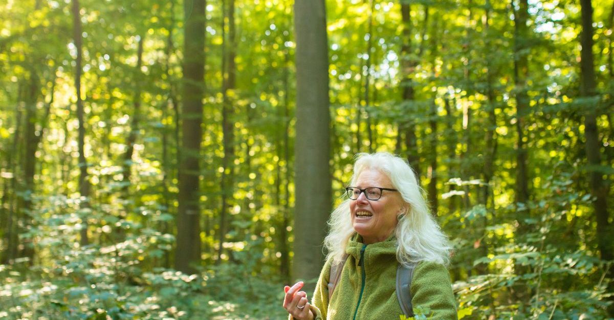 Eine Frau im Wald hält Wildkräuter in der Hand und trägt einen Korb.