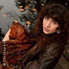 Eine Frau mit lockigem Haar sitzt in einem braunen Lederjacke am Wasser, umgeben von Herbstblättern.