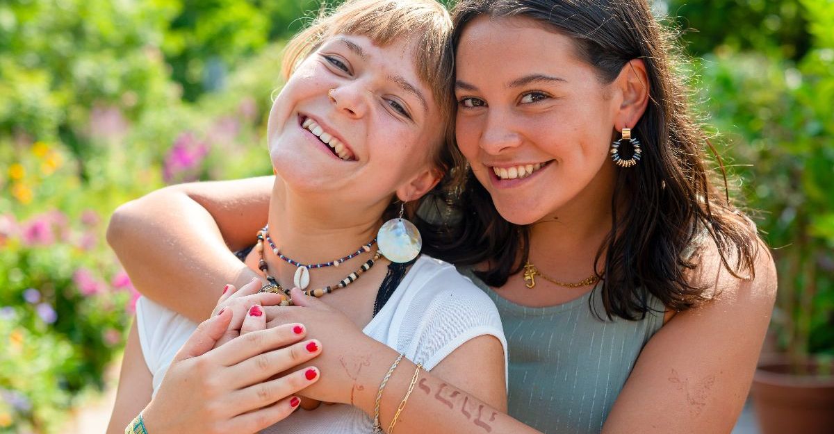Zwei junge Frauen umarmen sich lächelnd im Freien mit grüner Natur im Hintergrund.