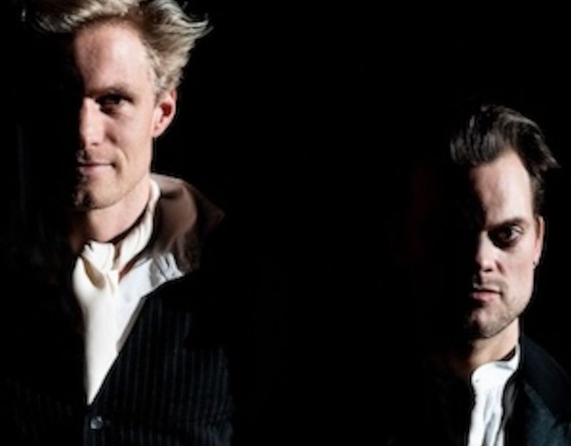 Zwei Männer in dunkler Beleuchtung, einer links mit hellem Haar, der andere rechts mit dunklem Haar, beide in formeller Kleidung.