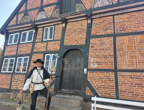 A historical timber-framed house with reddish-brown bricks and black beams. In front of the house stands a person in traditional clothing.