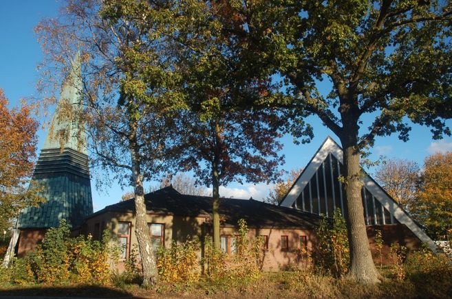 Kirche mit spitzem Turm und modernem Giebel, umgeben von Bäumen im Herbst.