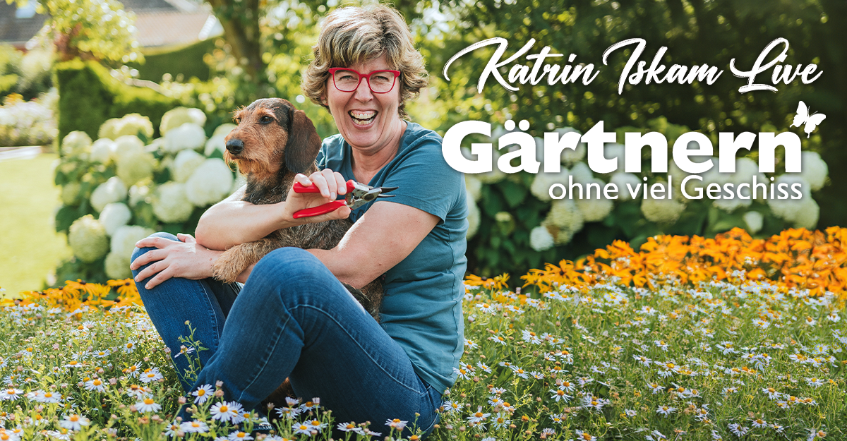 Eine Frau sitzt lächelnd mit einem Hund in einem blühenden Garten.