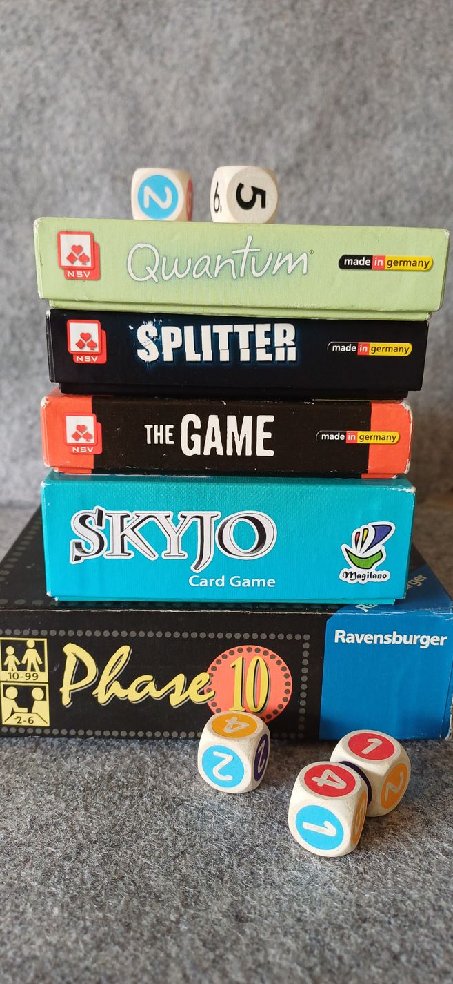 Ein Stapel von Brettspielen, darunter Qwantum, Splitter, The Game, Skyjo und Phase 10, mit mehreren bunten Würfeln davor.