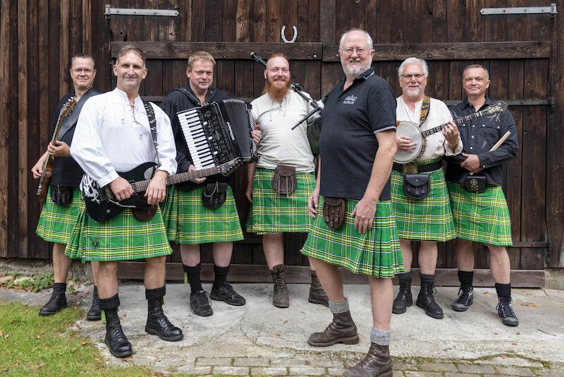 Eine Gruppe von sieben Männern in grünen Kilts posiert mit Musikinstrumenten vor einer Holzwand.