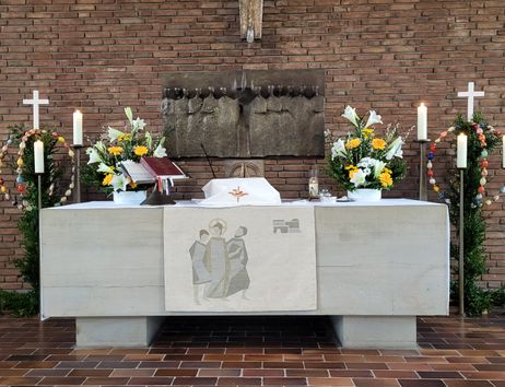 Kirchenaltar mit Blumen, Kerzen und einem Kreuz, umgeben von Ziegelwänden.