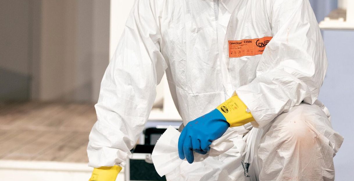 Person in Schutzanzug mit Maske und Handschuhen kniet auf einem Boden mit Reinigungsmaterial.
